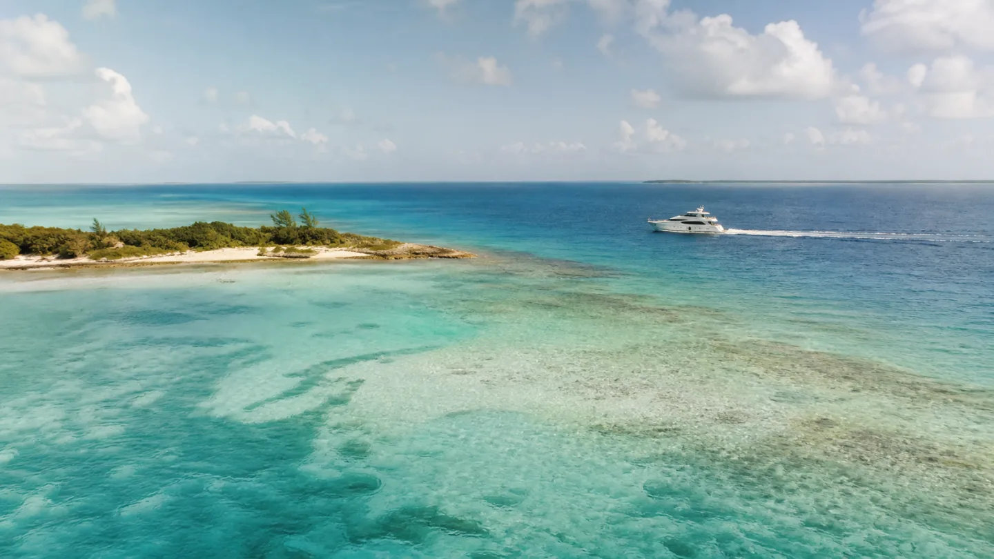 A flybridge yacht cruising beyond clear Bahamas shallows and a low island